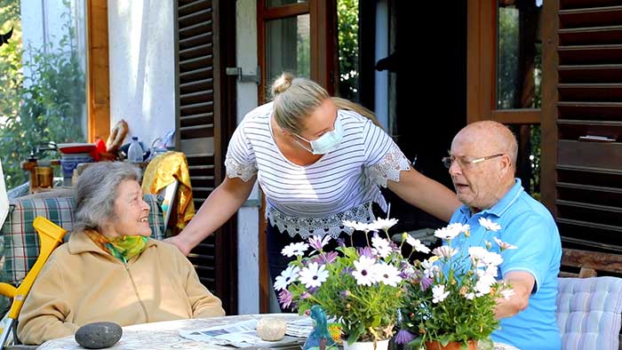Betreuerin mit Mundnasenmaske zwischen am Tisch sitzenden Seniorenpaar